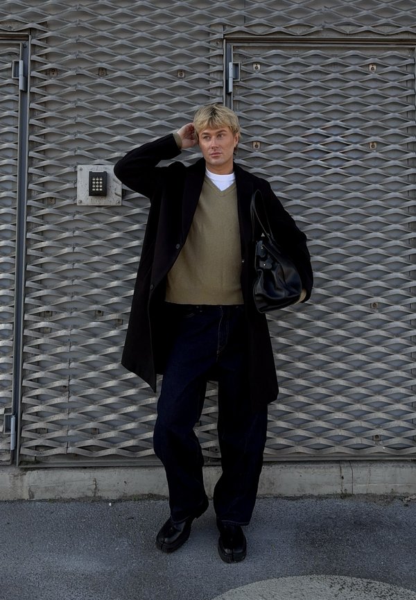 Una persona con indosso un cappotto nero sopra un maglione verde, jeans scuri e scarpe nere, che tiene in mano una borsa nera, è in piedi davanti a uno sfondo grigio testurizzato.