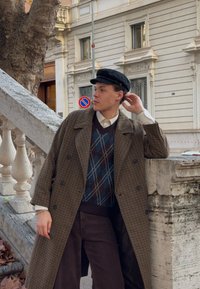 Brown houndstooth overcoat with large buttons, layered over a blue argyle jumper and dark trousers. Accessories include a black cap.
