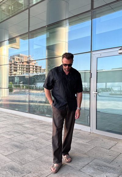Camisa corta de manga negra con una tela texturizada, pantalones rectos marrones y sandalias de tiras de color claro, de pie frente a puertas de vidrio.