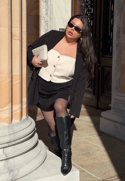 Mujer con blazer negro, top blanco abotonado, falda negra y botas negras altas, sosteniendo un clutch blanco cerca de columnas de piedra ornamentadas.