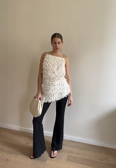 Mujer con top sin mangas blanco texturizado, pantalones negros de campana, sosteniendo un bolso de mano beige, de pie frente a una pared lisa beige.