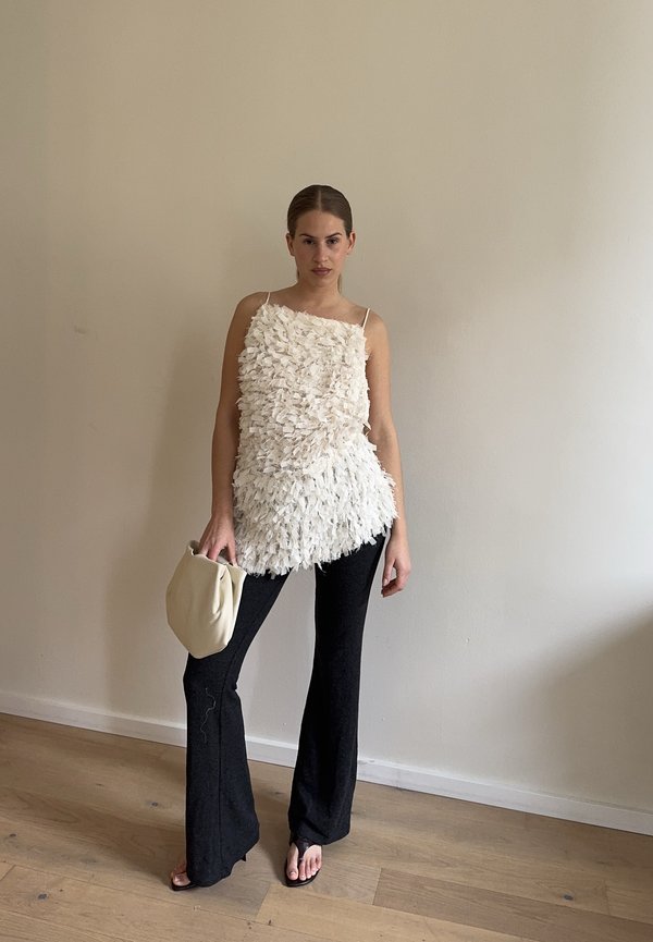 Γυναίκα που φοράει λευκό αμάνικο τοπ με υφή, μαύρο παντελόνι καμπάνα, κρατώντας ένα μπεζ τσαντάκι φάκελο, στέκεται μπροστά σε απλή μπεζ τοίχο.