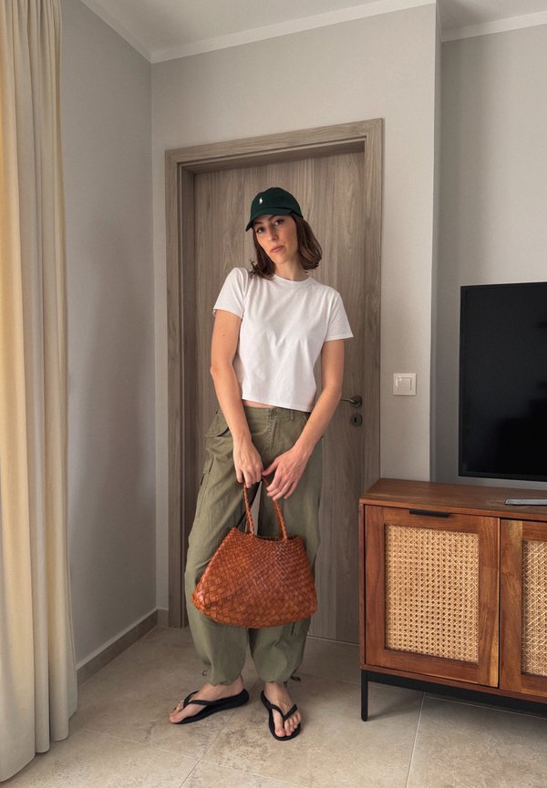 Femme en t-shirt court blanc, pantalon cargo vert, casquette noire et tongs, tenant un sac à main tressé marron dans une pièce moderne.