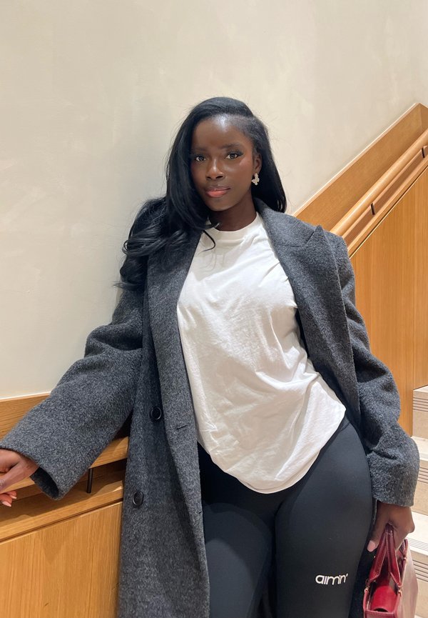 Femme aux longs cheveux noirs portant un manteau gris, une chemise blanche et des leggings noirs, debout à l'intérieur près d'un escalier en bois tenant un sac à main rouge.