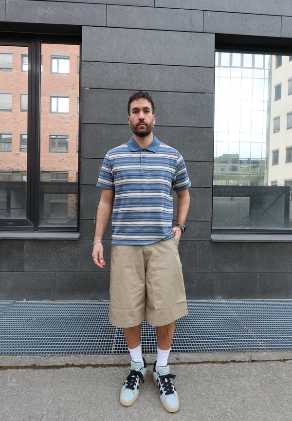 Mann som står mot en mørk flislagt vegg, iført en blåstripet piké, beige knelang shorts, hvite sokker og joggesko.