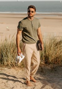 Camiseta henley verde, pantalones beige ajustados con puños elásticos, llevando sandalias blancas y una bolsa marrón, de pie en una playa de arena.