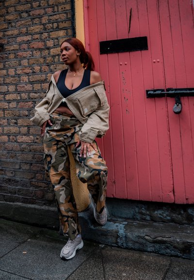 Chaqueta oversized de color beige claro, desabotonada, sobre un top corto negro, combinada con pantalones cargo de camuflaje. Zapatillas grises con suelas gruesas. Fondo de ladrillo.