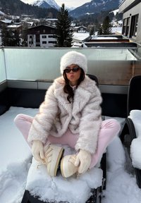 Plašč in klobuk iz umetnega krzna v svetlo bež barvi, rožnate nogavice, čevlji v krem barvi z vezalkami. V ozadju prizora sta sneg in gore.