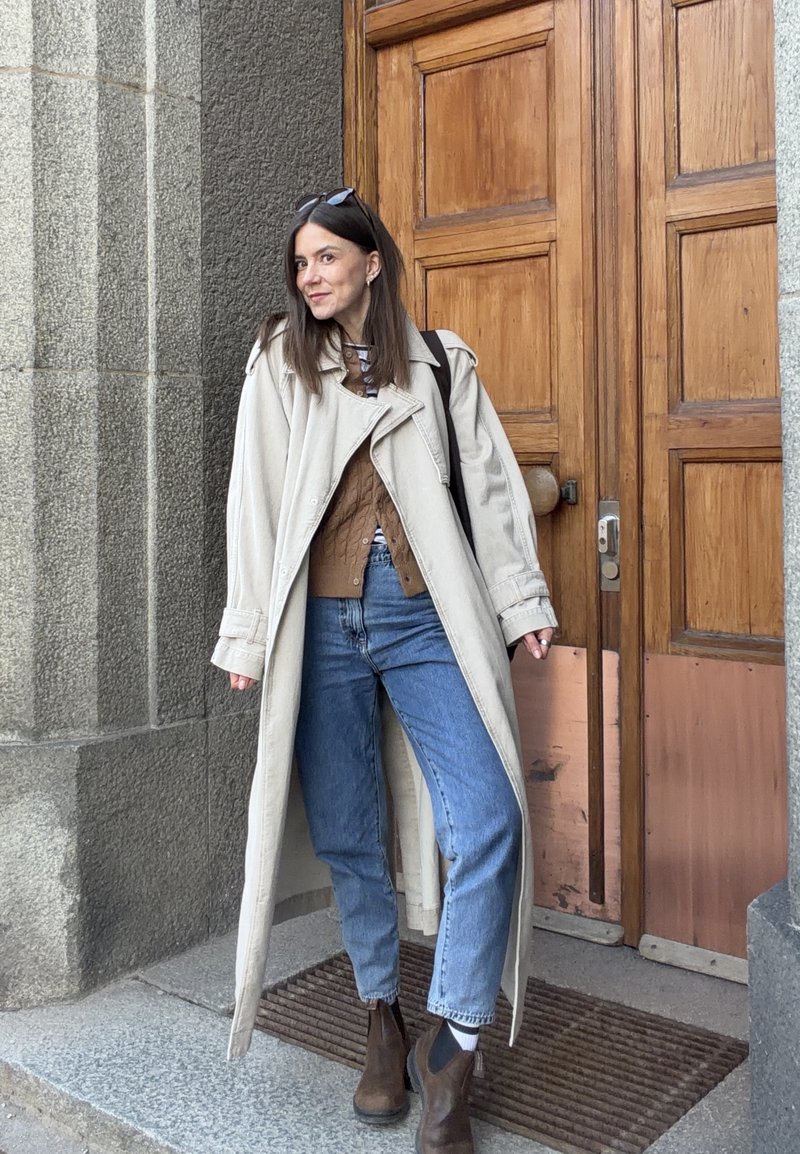 Jeune femme en long manteau beige, pull marron, jean bleu et bottes marron debout près d'une porte en bois.