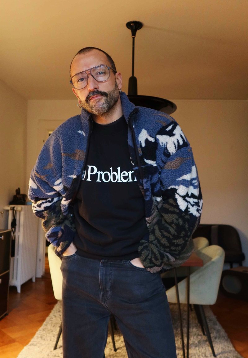 Homme chauve portant des lunettes et des boucles d'oreilles, vêtu d'une veste polaire à motifs et d'une chemise noire avec l'inscription « No Problem », debout dans une salle à manger chaleureuse.