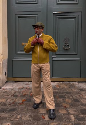 Hombre con chaqueta mostaza, guantes rojos, pantalones beige, zapatos negros y gorra verde, ajustando el cuello de la chaqueta en una calle de adoquines frente a grandes puertas grises.