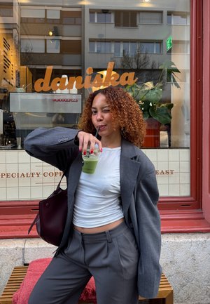 Mujer con cabello rizado, blazer gris y camisa blanca tomando una bebida verde con pajilla, guiñando un ojo frente a una cafetería llamada Daniska.