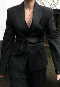 Blazer negro con solapas de satén, cintura ceñida, hombros estructurados y bolsillos frontales; combinado con una blusa negra. La tela tiene una textura suave.