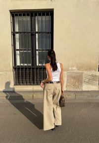 Bēša plašu kāju bikses ar aizmugurējiem kabatām, baltā ribotā krūštura krekls un mazs brūns rokassomu. Garas mati sasieti zirgaste. Sērsna uz ietves.