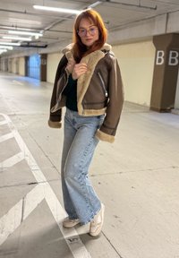 Veste courte en cuir marron avec col et poignets en fausse fourrure, associée à un jean évasé bleu clair et des baskets blanches. Cadre : parking intérieur.