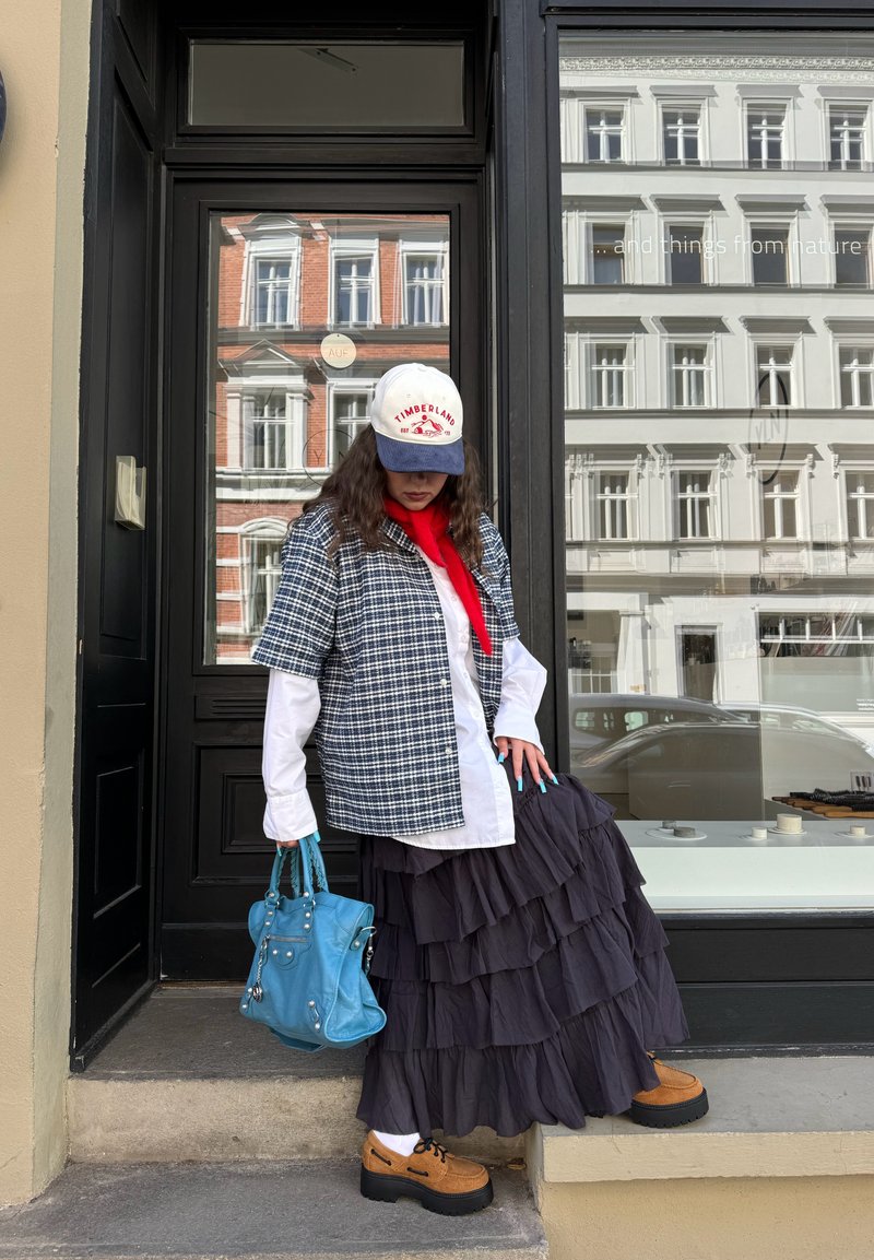Femme portant une jupe noire à couches, une veste à carreaux, une écharpe rouge et une casquette Timberland, tenant un sac à main bleu, debout près d'une porte noire.