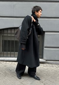 Μαύρο παλτό, μαύρο υπερμεγέθες παντελόνι και μαύρα παπούτσια. Κουβαλώντας μια σκοτεινή τσάντα με λουράκι. Ορατή υφή υφάσματος και πτυχές λεπτομέρειες.