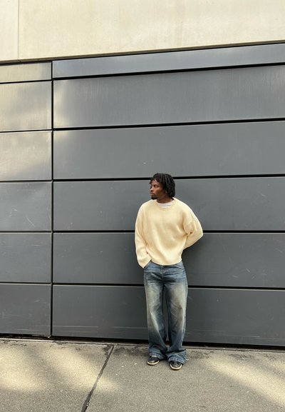 Suéter de punto beige claro, jeans de mezclilla azul y zapatos oscuros contra una pared gris texturizada; la figura está de pie con las manos en los bolsillos.