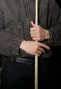 Braune Denimjacke mit gerafften Ärmeln, schwarzer Nieten-Gürtel und silbernen Ringen. Hände umklammern einen Holzstock; ein klarer Armreif ist sichtbar.