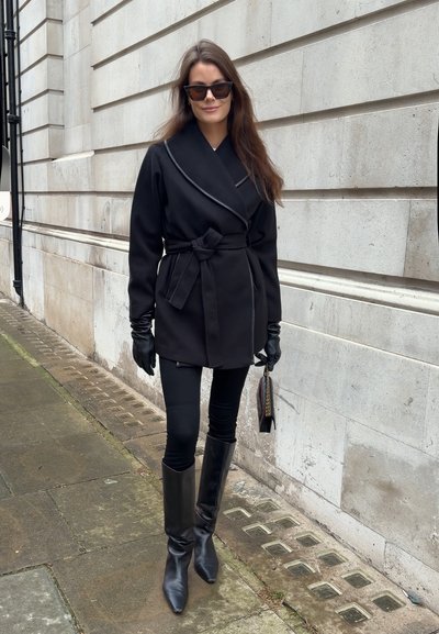 Mujer con abrigo negro con cinturón, guantes negros, botas, gafas de sol, sosteniendo un pequeño bolso negro, de pie en la acera junto a un muro de piedra.