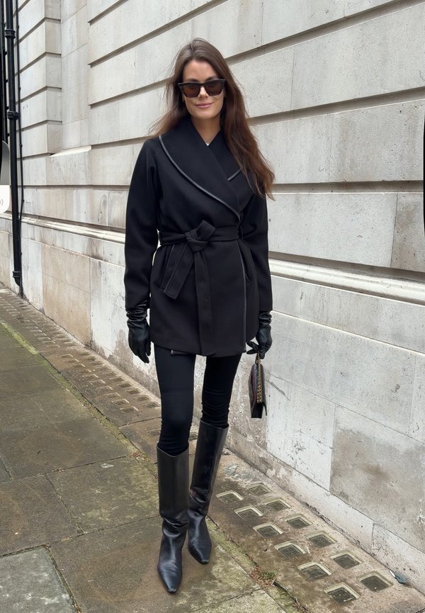 Žena u crnom kaputu s remenom, crnim rukavicama, čizmama i sunčanim naočalama, drži malu crnu torbicu, stoji na pločniku uz kameni zid.