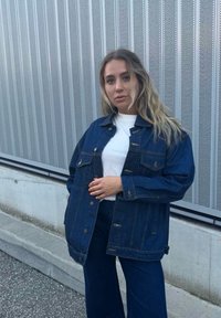 Τζάκετ τζιν σε σκούρο μπλε χρώμα με μεγάλα μπροστινά τσέπες, κλείσιμο με κουμπιά και χαλαρή εφαρμογή, συνδυασμένο με λευκό πουκάμισο και αντίστοιχο τζιν.