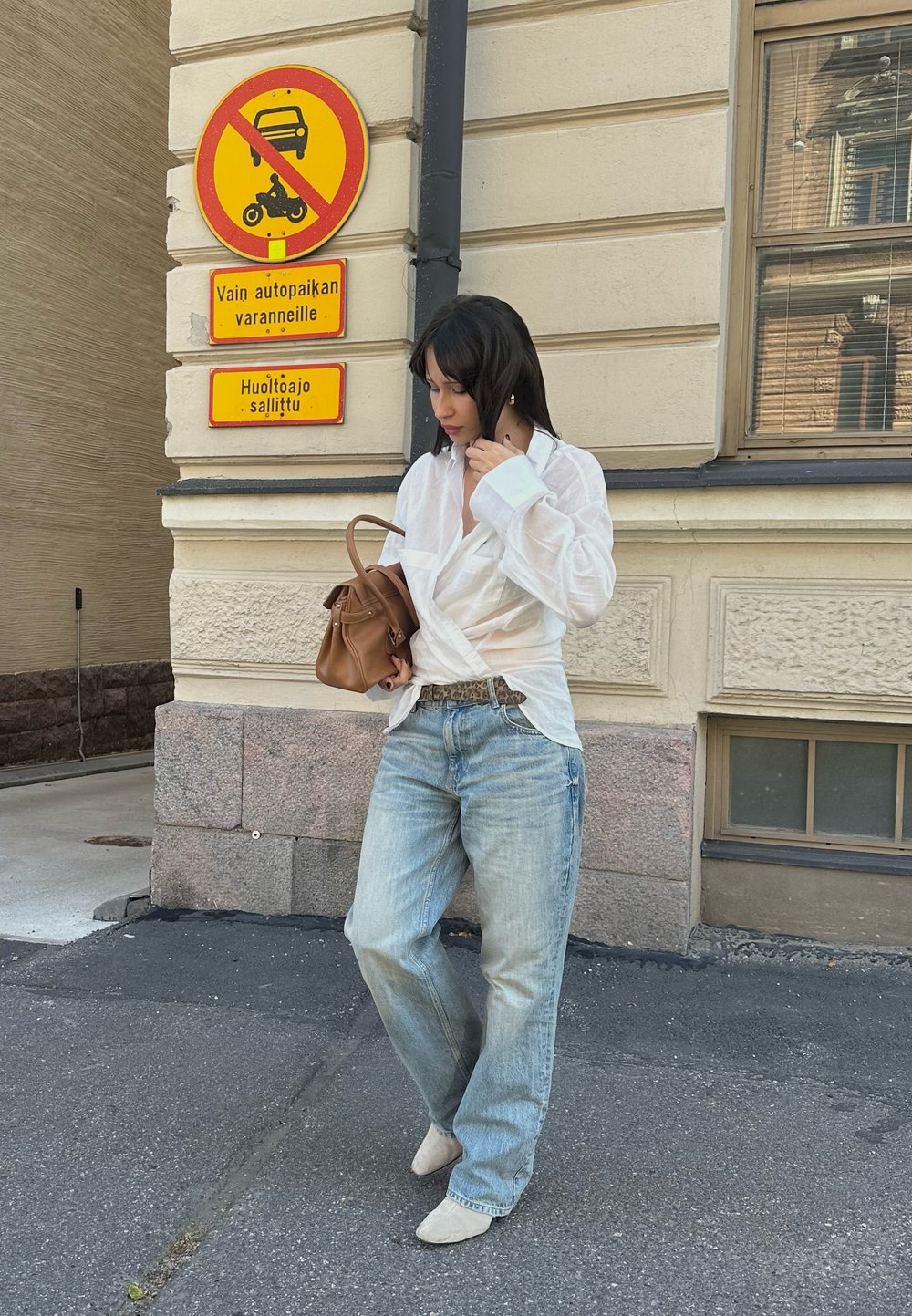 Witte wikkelshirt over lichtblauwe losse jeans, een bruin handtas dragend. Borden op de achtergrond geven parkeerbeperkingen aan. Stedelijke straatomgeving.