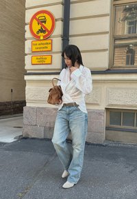 Witte wikkelshirt over lichtblauwe losse jeans, een bruin handtas dragend. Borden op de achtergrond geven parkeerbeperkingen aan. Stedelijke straatomgeving.