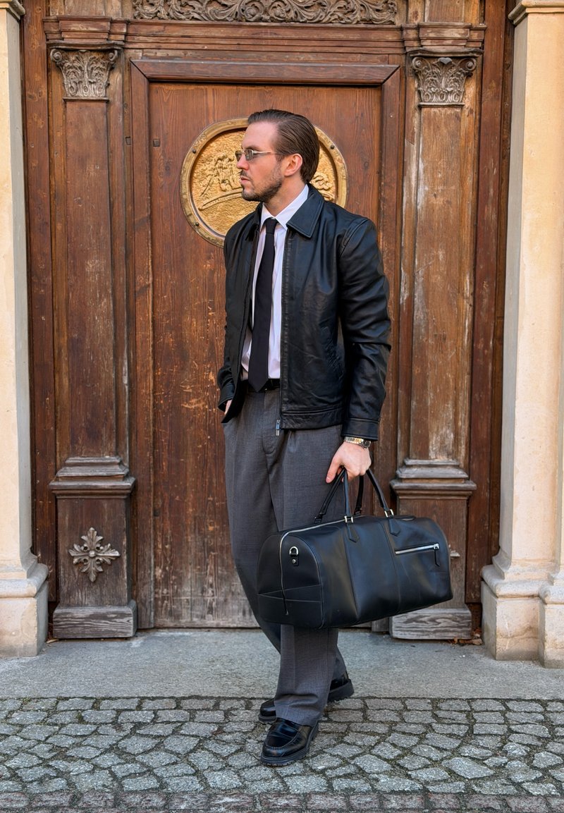 Homme en veste en cuir noire et cravate tenant un sac de sport noir, debout sur des pavés devant une porte en bois sculptée.