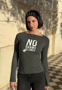 Femme avec coupe au carré noire et serre-tête, portant une chemise rayée à manches longues sur laquelle est écrit "NO DRAMA ALLOWED", debout sur un trottoir à damier.