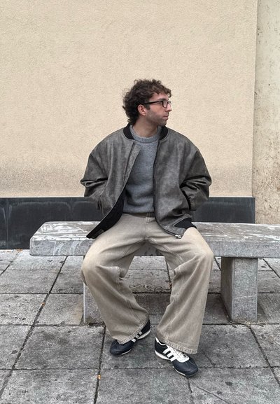 Chaqueta bomber gris con ribetes negros, suéter gris, pantalones beige holgados y zapatillas negras con acentos blancos, sentado en un banco de piedra.