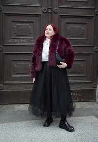 Γυναίκα με κόκκινα μαλλιά που φοράει μπορντό γούνινο μπουφάν, λευκό πουκάμισο, μακριά μαύρη φούστα τούλι και μαύρα platform παπούτσια με καρφιά, στέκεται δίπλα σε ξύλινη πόρτα.