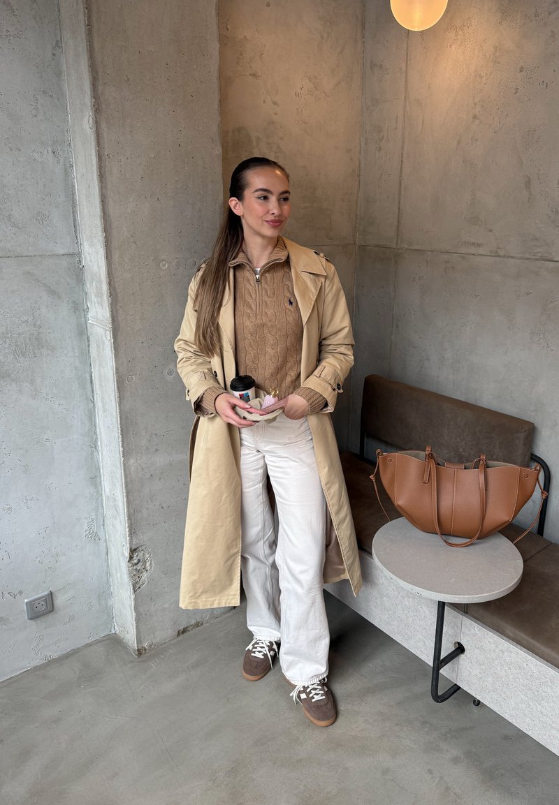 Trench coat bege, suéter de malha castanho, calças brancas de perna larga; ténis castanhos com atacadores brancos; bolsa castanha em cima de uma mesa cinza.