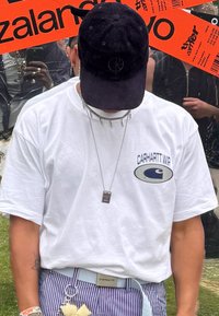 Biele tričko s modrou logom Carhartt, pruhované nohavice, modrý opasok, strieborný náhrdelník a čierna šnúrová čiapka s detailom loga.