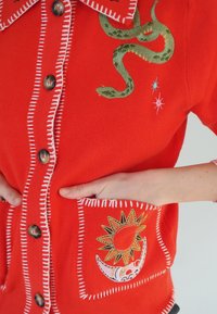 Cardigan rouge en maille avec broderie de serpent verte, coutures blanches, grands boutons et poche à motif représentant un soleil et un croissant de lune.