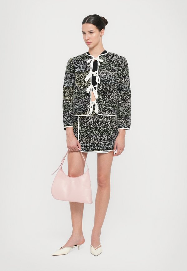 Schwarz-weiße gemusterte Jacke mit Spitzenbindebändern, kombiniert mit einem passenden Rock. Das Model hält eine sanft rosa Lederhandtasche und trägt beige Absätze.