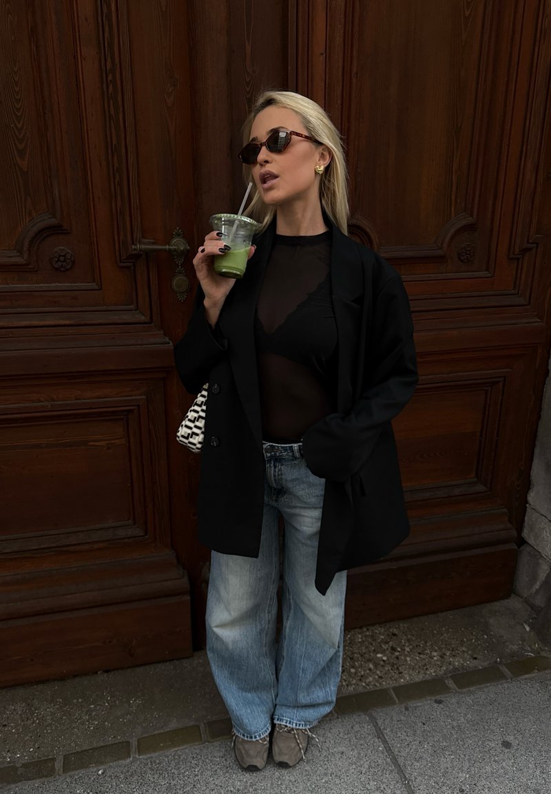 Femme portant des lunettes de soleil, un blazer noir et un top transparent, un jean large, tenant un smoothie vert avec une paille, debout près d'une grande porte en bois.