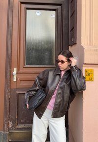 Dark brown leather jacket, pink sweater, white trousers, and a black woven clutch. The setting includes a brown door with a glass panel.
