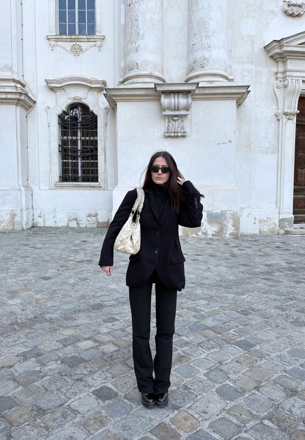 Nainen pukeutuneena mustaan pukuun ja aurinkolaseihin, kantaa valkoista käsilaukkua ja seisoo mukulakivikadulla koristeellisen valkoisen rakennuksen julkisivun edessä.