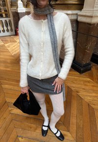 Femme debout à l'intérieur sur un sol en bois portant un pull blanc duveteux, une jupe grise, des collants blancs à motif, des chaussures noires, une écharpe grise, tenant un sac à main noir.