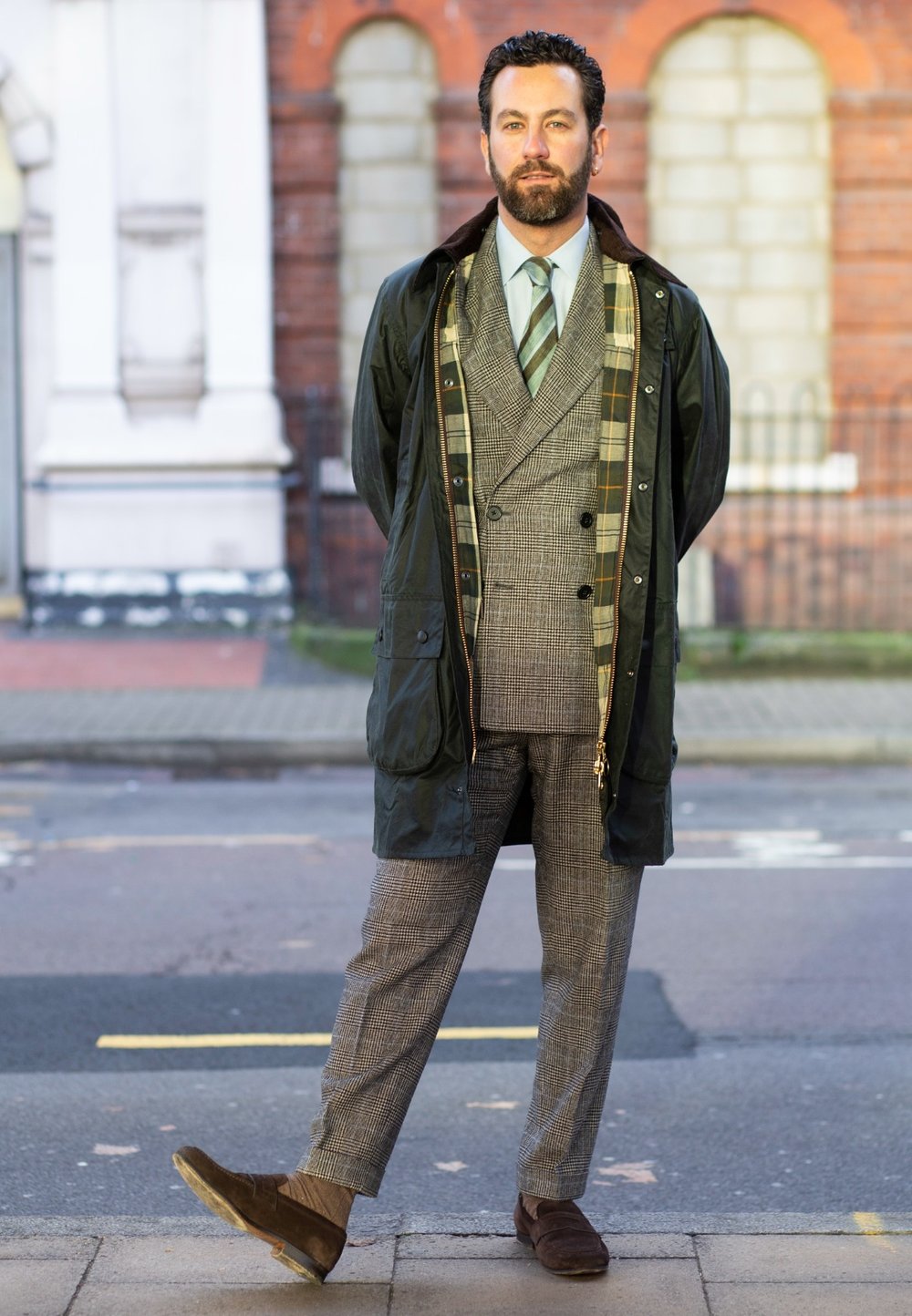 Green waterproof jacket worn over a grey checked suit, complemented by a light blue shirt and a striped tie, along with brown suede shoes.
