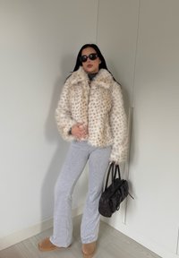 Manteau en fausse fourrure avec un motif tacheté crème et marron, associé à un pantalon évasé gris et des bottines à enfiler couleur tan, tenant un sac à main noir.