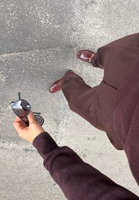 Burgundowy sweter, pasujące spodnie i burgundowe mokasyny. Trzyma czarne okulary przeciwsłoneczne w prostokątnej oprawie i ciemnych szkłach.