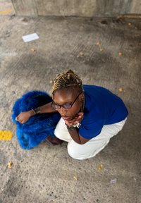 Modell in blauem Hemd und weißer Hose, sitzt neben einem blauen Kunstpelzobjekt, posiert mit geflochtenen Haaren und übergroßen Brillen.