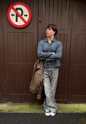 Joven con pelo rizado, vistiendo camisa de cuadros y vaqueros grises, se apoya en una puerta marrón bajo una señal de prohibido estacionar, sosteniendo una chaqueta marrón.
