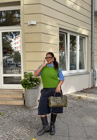 Zielona dziergana kamizelka, niebieska w paski koszula, czarne szerokie szorty, czarne wysokie buty do kolan i oliwkowa torebka z gwiazdkowym akcentem. W pobliżu doniczkowa roślina.