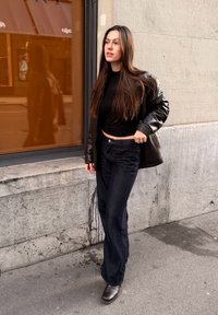 Melna ādas jaka pār piegulošu melnu topu, kombinēta ar tumšām džinsu biksēm un brūnām potīšu zābakiem, stāvot pie stikla loga ar oranžu atspulgu.