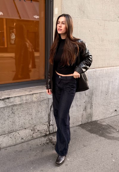 Chaqueta de cuero negra sobre una camiseta negra ajustada, combinada con jeans oscuros y botines marrones, de pie cerca de una ventana de vidrio con un reflejo naranja.