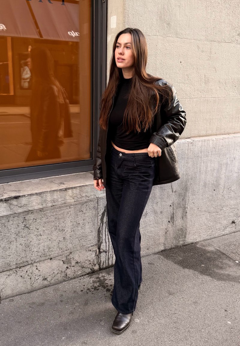 Giacca di pelle nera su una maglietta nera aderente, abbinata a jeans scuri e stivaletti marroni, in piedi vicino a una finestra di vetro con un riflesso arancione.