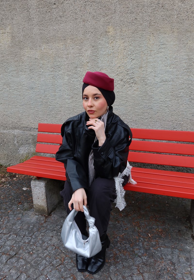 Mujer con boina roja y chaqueta de cuero negra sentada en un banco rojo al aire libre, sosteniendo un bolso plateado sobre un suelo de adoquines.